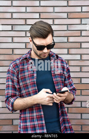 Vorderansicht des gutaussehenden jungen Mann in Smart Casual Wear halten ein Handy im Stehen an der Wand im Hintergrund. Stockfoto