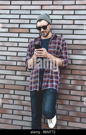 Hipster sms Phone App in Stadt Straße Mauer Hintergrund. Erstaunlicher Mann holding Smartphone in Smart Casual Wear stehen. Urban young profe Stockfoto