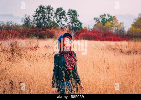 Tolle Frau im Park spazieren geht in trüben Wetter im Herbst. Rothaarige Frau in einem blauen Hut, Mantel und Sonnenbrille geht das Feld bei Fallen suchen Stockfoto