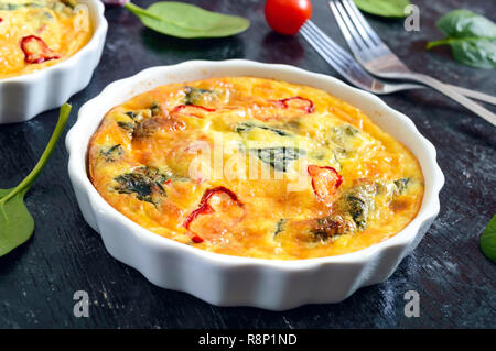 Frittata mit frischem Gemüse und Spinat. Italienische Omelette in keramischen Formen auf einem schwarzen Hintergrund. Stockfoto