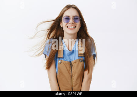 Taille-up Shot von charmanten Sorglos kaukasischen Freundin mit langen braunen Haaren flippen auf Luft zu lächeln und zu lachen freundlich und spielerisch posiert vor grauem Hintergrund in der braunen Overalls und Sonnenbrille Stockfoto