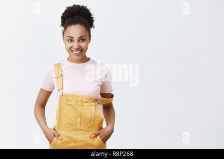 Lassen Sie uns ändern Welt beginnen. Portrait von charmanten zuversichtlich und fröhlich Afrikanische amerikanische Frau in gelber Latzhose, mit Gurt auf der Schulter, halten die Hände in den Taschen und breit grinsend, bereit für Aktion Stockfoto