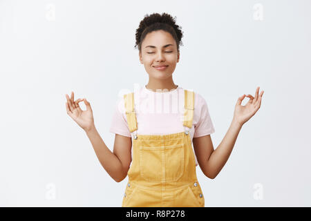 Beruhigung und Beseitigen von Stress im Inneren. Ruhige und nette gut aussehende dunkelhäutige Frau im gelben trendigen Overall, halten sich an den Händen an beiden Seiten mit Zen Geste, Meditation oder Yoga Stockfoto