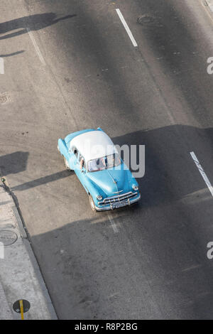 Aus der Vogelperspektive eines blauen Oldtimers aus den 1950er Jahren, das auf einer sonnendurchfluteten Asphaltstraße in einer urbanen Umgebung mit minimalem Verkehr in Havanna Kuba fährt Stockfoto