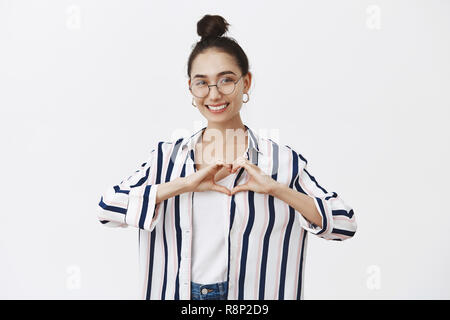 Portrait von attraktiven Angebot feminin Freundin mit bun Frisur in der stilvollen Kleidung und Gläser Gestaltung Herz mit Hände über Brust und breit grinsend, während Liebe und Sympathie Stockfoto