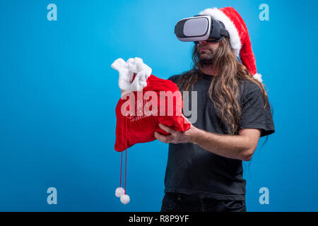 Portrait der männlichen tragen Santa Claus Hut, Tasche und das Tragen eines VR-Gang in der Landschaft. Studio shot vor blauem Hintergrund Stockfoto
