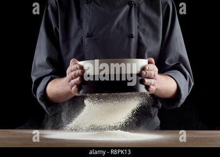 Männliche Hände sichten Mehl aus alte Sieb auf alten hölzernen Küchentisch. Auf schwarzem Hintergrund isoliert. Stockfoto