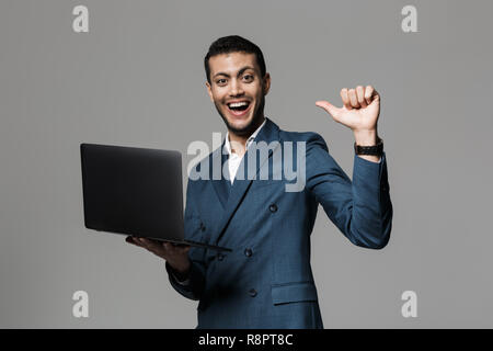 Bild des erfolgreichen arabischer Geschäftsmann 30 s in Anzug und lächelnd Laptop isoliert über grauer Hintergrund Stockfoto