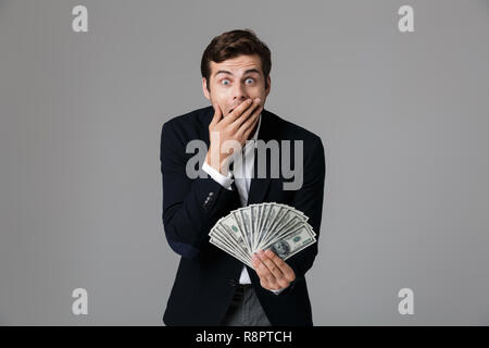Bild von überrascht Geschäftsmann 30 s in Anzug und lächelnd Fan von Geld in Dollar Banknoten über grauer Hintergrund Stockfoto