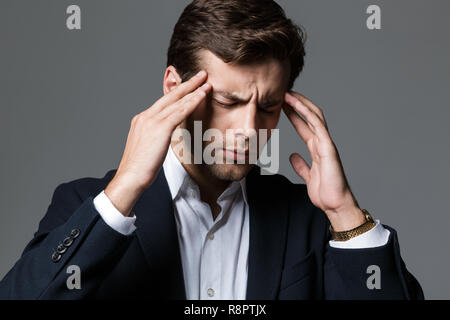 Nahaufnahme, Porträt einer erschöpften jungen Geschäftsmann in Anzug über grauen Hintergrund isoliert gekleidet, leiden unter Kopfschmerzen Stockfoto
