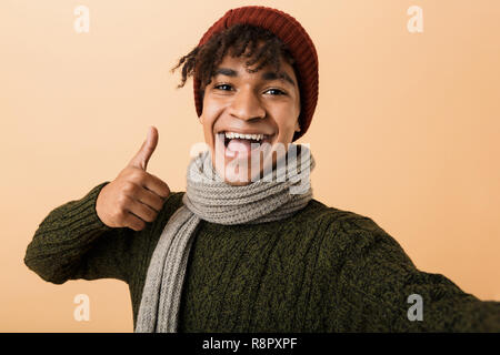 Portrait von Happy Teen Mann mit Hut und Schal gestikulierend an Kamera über Gelb Hintergrund isoliert Stockfoto