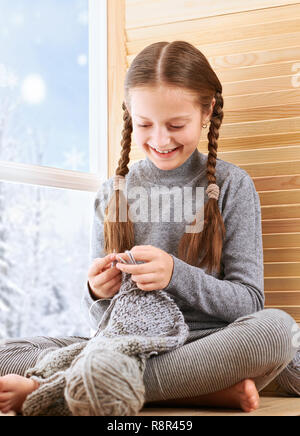 Kind, Mädchen sitzt auf dem Fensterbrett und das Stricken der Schal aus grauer Wolle Garn. Schöne Aussicht aus dem Fenster-sonnigen Tag im Winter und Schnee. Stockfoto