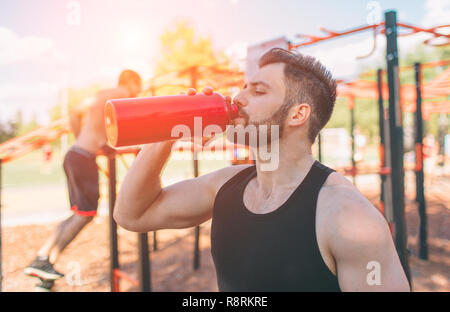 Mann mit Bügeleisen Flasche und Trinkwasser. Junge schöne gut geformt, sportlichen Mann Ausruhen nach dem Training im Freien. Männliche fitness Modell Training Training. Stockfoto