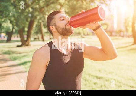 Mann mit Bügeleisen Flasche und Trinkwasser. Junge schöne gut geformt, sportlichen Mann Ausruhen nach dem Training im Freien. Männliche fitness Modell Training Workout Stockfoto