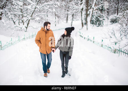 Glückliches junges Paar im Winter. Familie im Freien. Mann und Frau auf der Suche nach oben und Lachen. Liebe, Spaß, Jahreszeit und Personen - Wandern im Winter Park. Stand und halten einander die Hände Stockfoto