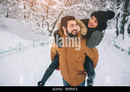 Glückliches junges Paar im Winter. Familie im Freien. Mann und Frau auf der Suche nach oben und Lachen. Liebe, Spaß, Jahreszeit und Personen - Wandern im Winter Park. Stand und halten einander die Hände. Sie auf seinem Rücken. Stockfoto