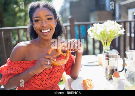 Porträt Lächeln, selbstbewussten jungen Frau mit Brezel geniessen Sie das Frühstück auf der sonnigen Balkon Stockfoto