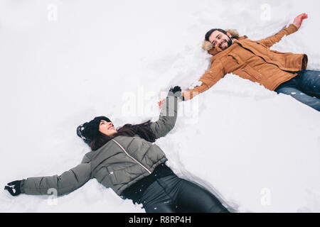 Glückliches junges Paar im Winter. Familie im Freien. Mann und Frau auf der Suche nach oben und Lachen. Liebe, Spaß, Jahreszeit und Personen - Wandern im Winter Park. Liegen im frischen Schnee, Schnee Engel Spaß Stockfoto