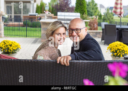 Fett Mann in Gläsern und schwarzen Pulli und Frau mit kurzen blonden Haaren sitzen auf einem Sofa, zurück in die Kamera schauen und lächeln, sitzen auf den offenen Terr Stockfoto
