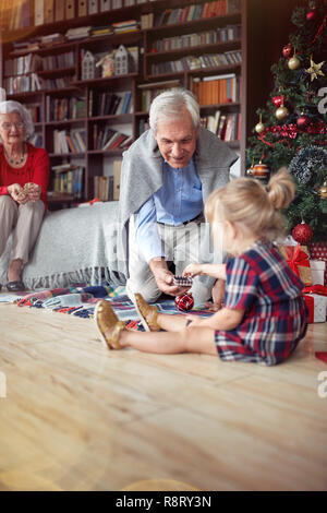Nettes Mädchen ist offen Weihnachtsgeschenk vor einem eingerichteten X-mas Tree mit Großvater Stockfoto