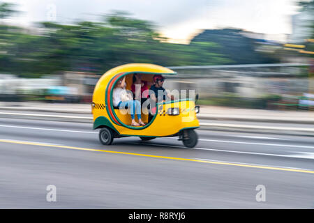 Kubanische Mann, der eine Beschleunigung helle gelbe Coco Taxi mit zwei Passagiere in Havanna, Kuba Stockfoto