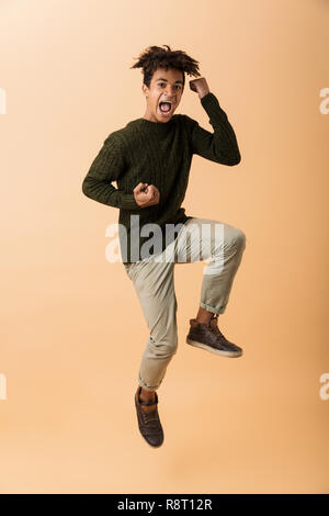 Volle Länge Foto von Teenage african american Guy tragen Pullover lachen und springen über beigen Hintergrund isoliert Stockfoto