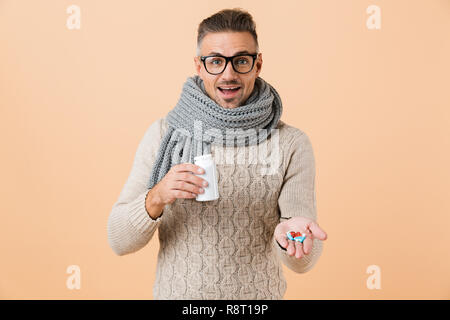 Porträt von einem freundlichen Mann in Pulli und Schal stehend über beigen Hintergrund isoliert gekleidet, mit Pillen Kapseln Stockfoto