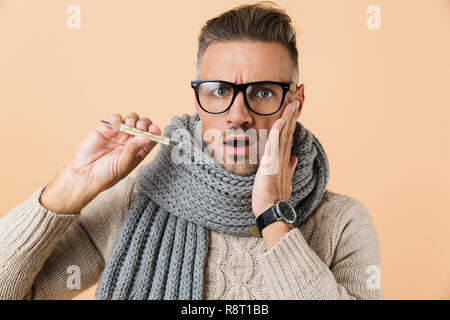 Porträt eines kranken Mann in Pulli und Schal stehend über beigen Hintergrund isoliert gekleidet, mit Thermometer Stockfoto