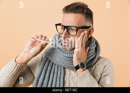 Porträt von einem besorgten kranken Mann in Pulli und Schal stehend über beigen Hintergrund isoliert gekleidet, mit Thermometer Stockfoto