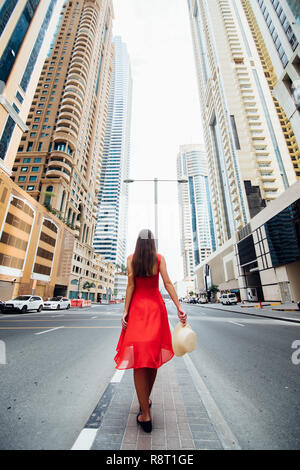 Junge Frau im roten Kleid und Strohhut zu Fuß in der Nähe von Wolkenkratzern in der modernen Stadt. Low Angle View Stockfoto