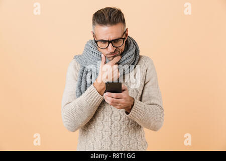 Porträt einer nachdenklichen Mann in Pulli und Schal stehend über beigen Hintergrund isoliert gekleidet, mit Handy Stockfoto