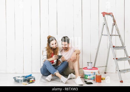 Foto von nettes Paar Mann und Frau sitzt auf dem Boden über weiße Wand in der Nähe der Leiter und über Handy während der Renovierung innen Stockfoto