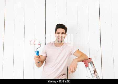 Foto von brünett Mann 20 s Malerei weiße Wand und mit Paint roller Renovierung Stockfoto