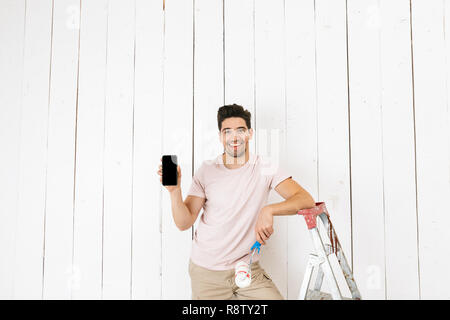 Foto von schöner Mann 20 s mit Handy beim Malen weiße Wand mit farbroller und Renovierung Stockfoto