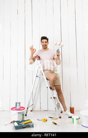 Volle Länge Foto von fröhlicher Kerl 20 s auf Leiter und Malerei Wand während der Sanierung stehen Stockfoto
