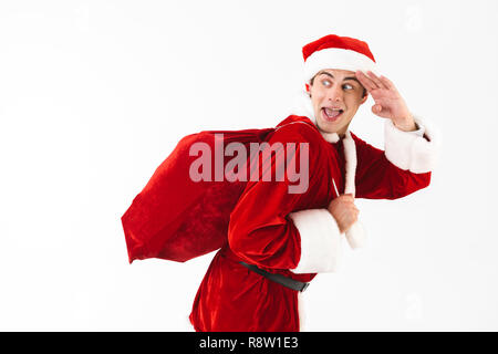 Portrait von optimistischen Mann 30 s in santa claus Kostüm und Red Hat mit Geschenk Tasche über der Schulter auf weißem Hintergrund im Studio isoliert Stockfoto