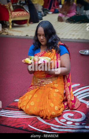 Ein frommer Hindu Frau in einem schönen orange Sari kniet im Gebet & vor Statuen von Gottheiten, während eine Nahrung bietet. In New York City Stockfoto