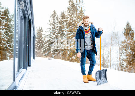 Porträt eines Mannes im Winter Kleidung stehend mit Schneeschaufel auf der Terrasse des Gebäudes in den Bergen Stockfoto