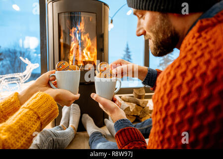 Junges Paar in hellen Pullover mit heißen Getränken in der Nähe der Feuerstelle im modernen Haus in den Bergen gekleidet durnig Winter Stockfoto