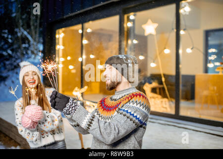 Junge glückliches Paar in Pullover feiern Winterurlaub mit bengalischen Feuer vor einem wunderschönen eingerichteten Haus gekleidet Stockfoto