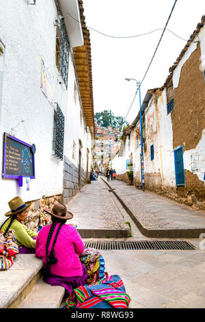 Quechua peruanische Frauen sitzen auf dem Straßenpflaster einer kleinen Straße in San Blas und tragen helle Kleidung, Hüte und geflochtene Haare, Cusco, Peru Stockfoto