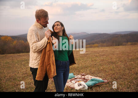 Zwei Menschen, junges Paar, 20-29 Jahre, schöne Geste Gentleman setzt seine Jacke auf Frau zurück und stand hinter ihr. Picknick im Freien auf Berg f Stockfoto
