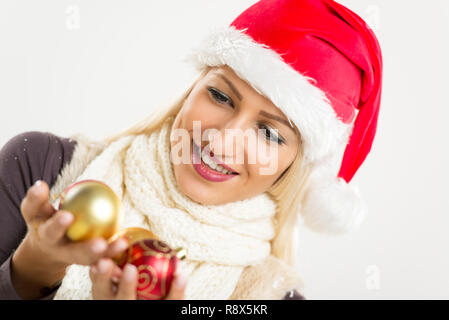 Süße blonde Mädchen mit Santa Claus hat und einen warmen Schal um den Hals, die eine dekorative Kugel mit einem Lächeln auf ihrem Gesicht. Stockfoto