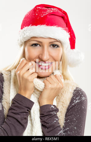 Junge schöne Blondine mit einem Santa Mütze auf dem Kopf, mit Schnee bedeckt, Lächeln sieht in die Kamera und stopfte es in ihr Schal. Stockfoto