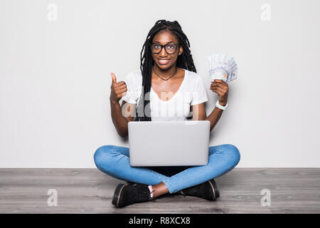 In voller Länge Porträt des jungen Afro-amerikanische Frau sitzt auf dem Boden halten Geld pc Ion grauer Hintergrund Stockfoto