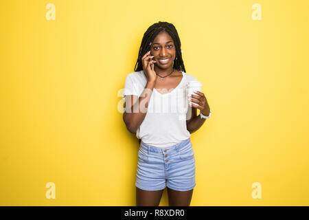 Attraktive dunkelhäutige Mädchen mit Afro Frisur tragen weiße T-Shirt in Telefongespräch mit ihrem Freund und trinken Kaffee isoliert auf Gelb Stockfoto