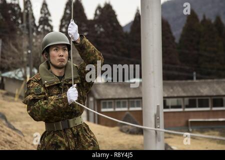 Ein service Mitglied mit der japanischen Boden Verteidigung-kraft die japanische Flagge während der Morgen Farben in der Hijudai Manöver, Japan wirft, 6. März 2017. Us-Marines und Matrosen an der Artillerie relocation Training Programm rechtzeitige und genaue Brände zu stellen militärische beruflichen Spezialgebiet Fähigkeiten, Zug Marines/Matrosen in gemeinsamen Fähigkeiten zu erhalten, und professionelle militärische Ausbildung für das allgemeine Ziel der Verbesserung der Bekämpfung der Betriebsbereitschaft und internationale Beziehungen zu fördern. Stockfoto