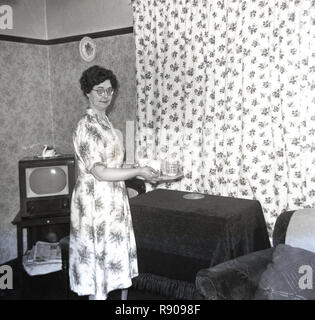 1940, historische, eine Dame in einem gemusterten Kleid in Ihrem Zimmer vorne, ein Tablett mit Tee Tassen und Gläser auf einem kleinen Tisch. Ein Fernseher der Ära ist in der Ecke des Raumes, England, UK gesehen. Stockfoto