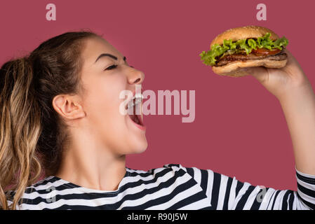 Crazy hungrig cool Girl in gestreiften T-Shirt essen Hamburger im Profil über bunte rosa Hintergrund Stockfoto