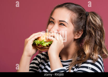 Crazy Lustig Cool girl in gestreiften T-Shirt essen Hamburger mit Appetit über bunte rosa Hintergrund Stockfoto